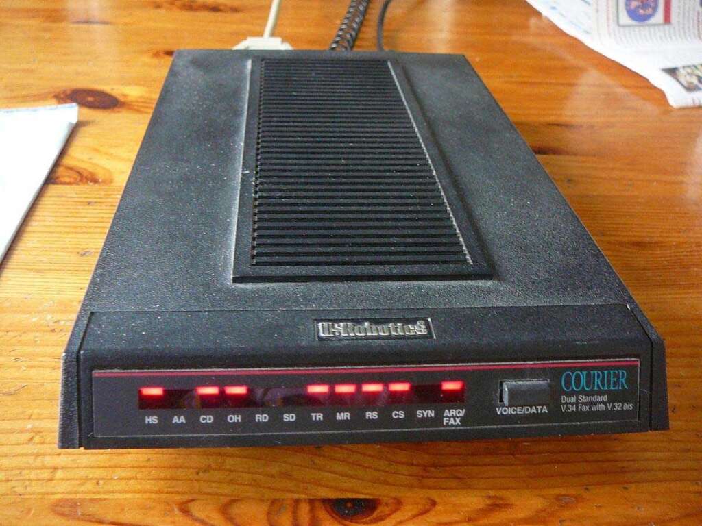 A vintage US Robotics Courier Dual Standard modem on a wooden surface, with multiple red indicator lights illuminated on its front panel. A power cable is connected at the back. A newspaper can be seen in the background.