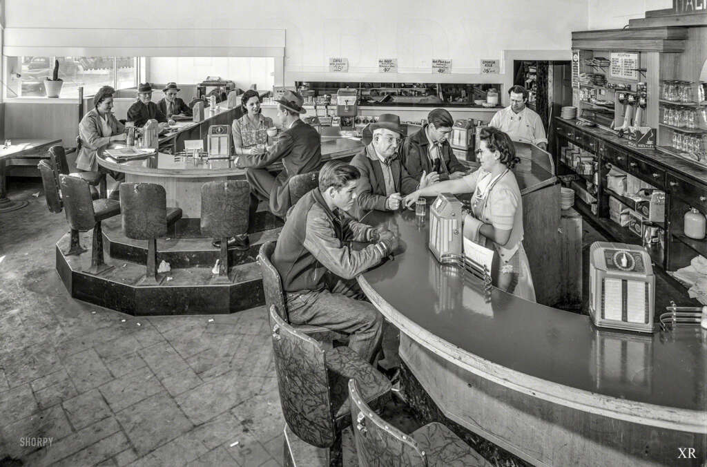 Black and white photo of a bustling diner with customers sitting at counters. A waitress serves a drink to a man. The room has a vintage look, with signs on the walls and retro furnishings, capturing a lively atmosphere.