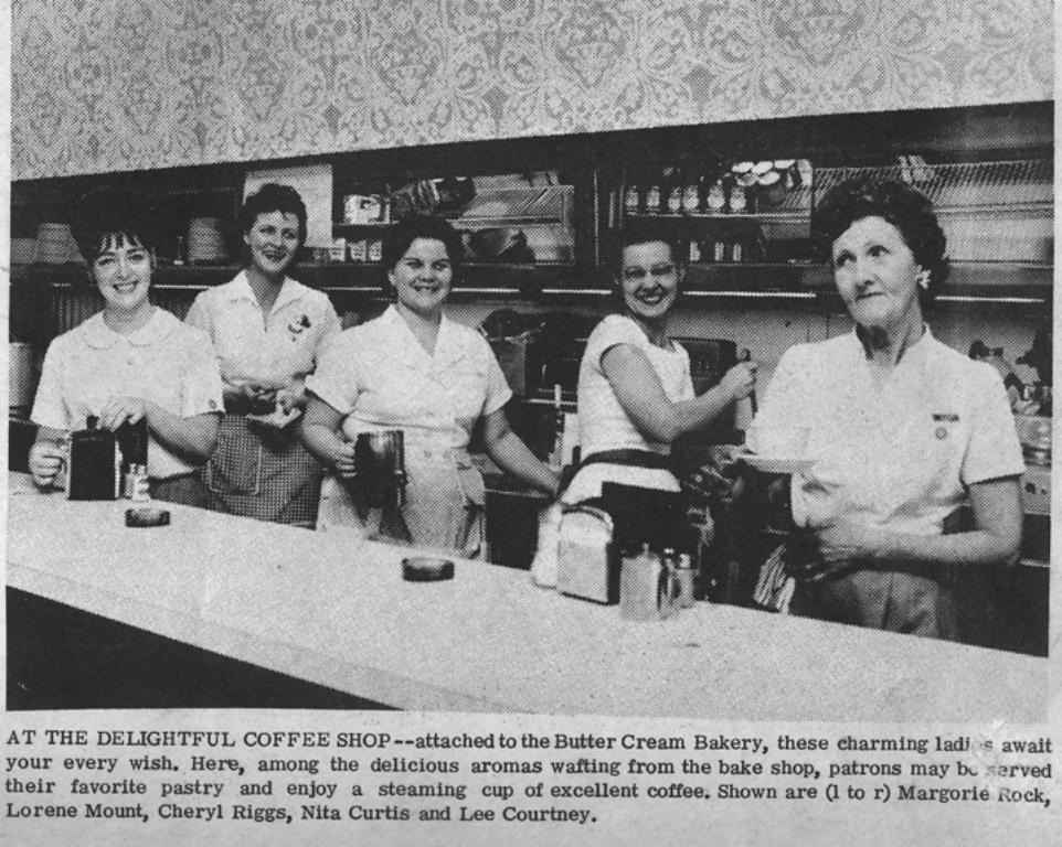 A vintage photo of five women standing behind a counter at a coffee shop. They are smiling, dressed in uniforms, surrounded by coffee pots and cups, with a mixer visible. A newspaper caption introduces them as staff at the Butter Cream Bakery.