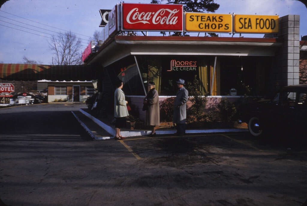 Three people stand outside a retro diner with signs for Coca-Cola, steaks, chops, and seafood. The diner has a "Moore's Ice Cream" sign, and there is a parked car nearby. The setting appears to be from the mid-20th century.