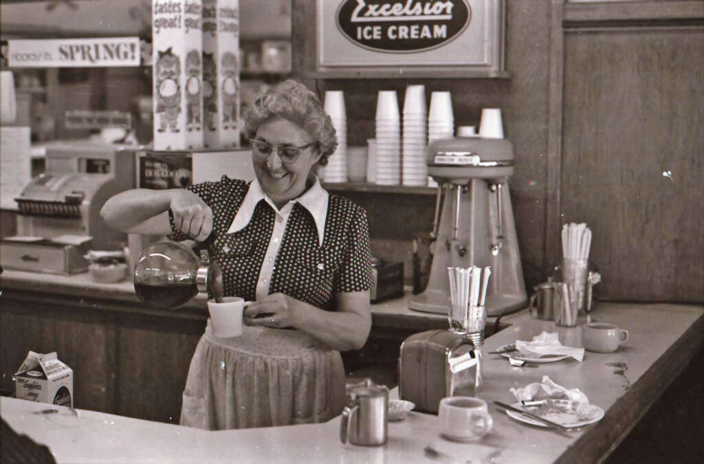 A smiling woman in a polka dot dress and apron pours coffee behind a diner counter. A vintage "Excelsior Ice Cream" sign hangs on the wall, and various kitchen supplies and appliances are visible. Cups and a milk carton are on the counter.