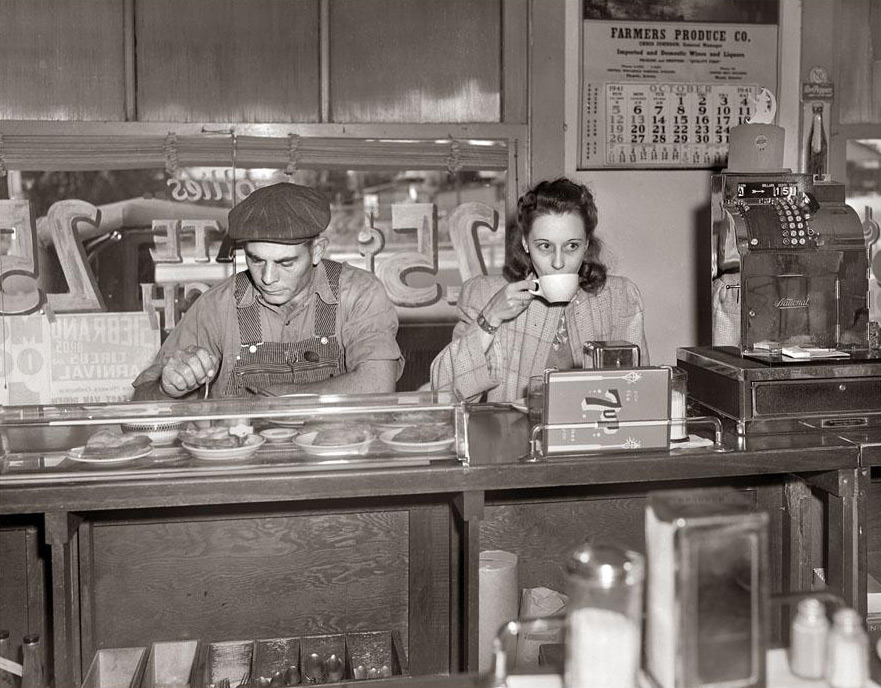 A man and woman sit at a diner counter. The man wears a cap and overalls, looking down at a plate of food. The woman sips from a cup, holding it with both hands. A cash register and a calendar are visible in the background.