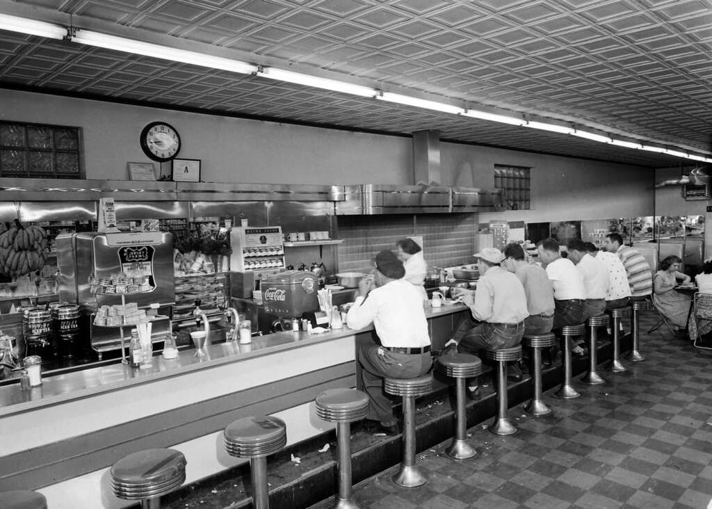 A vintage diner with a long counter and cushioned stools. Several people sit at the counter, facing a wall with a clock and menu board. Behind the counter, there are soda machines and various items for food preparation. The ceiling has a patterned design.