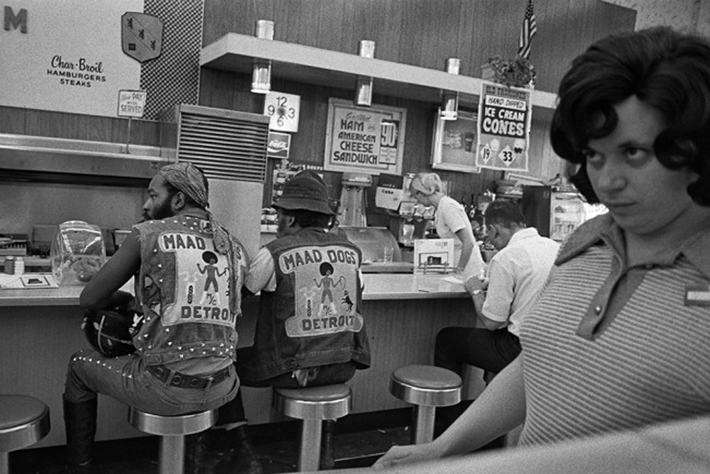 Two men wearing "Maad Dog" biker vests sit at a diner counter. Signs advertise items like hot dogs and ice cream cones. A woman with dark curly hair and a striped shirt stands in the foreground, looking to the side.