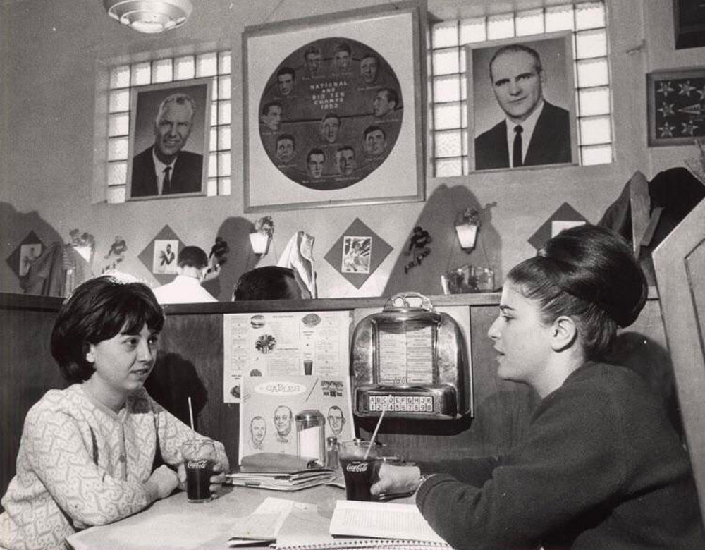 Two women sit in a vintage diner booth, each holding drinks. A jukebox is on the table. Behind them are framed portraits and a collage. The decor includes geometric wall patterns, contributing to a classic mid-20th-century ambiance.