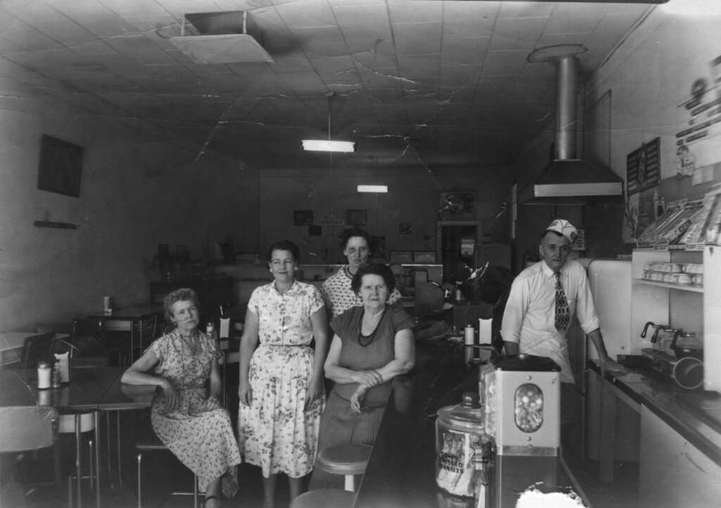 Five people are in a vintage diner setting. Four women in patterned dresses sit and stand in the foreground, while a man in a white uniform stands near a counter with kitchen equipment in the background. The room has a nostalgic atmosphere.