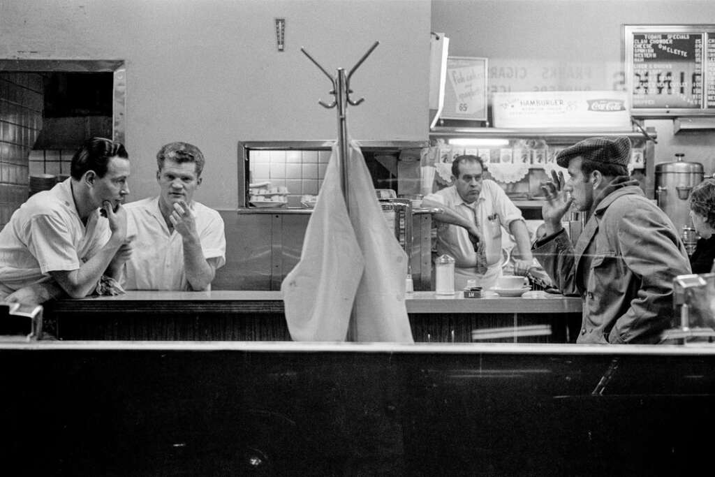 Black and white photo of a diner scene. Two men in white uniforms stand behind the counter, while two patrons sit opposite. A jacket hangs on a coat rack in the foreground. The setting is casual, with signs and a lunch menu visible.