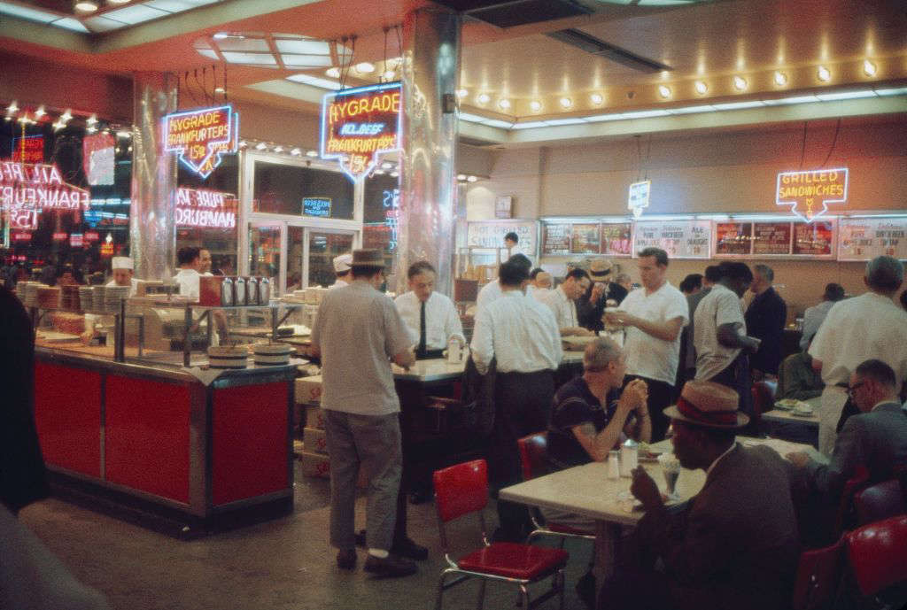 A busy 1950s-style diner with people eating at tables and standing at the counter. Neon signs advertise "HYGRADE" and "SANDWICHES," and red chairs and vintage decor contribute to the retro atmosphere.