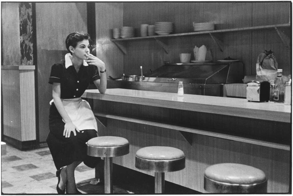 Black and white photo of a woman in a waitress uniform sitting on a stool at a diner counter. She is smoking a cigarette, with empty stools beside her. Plates and condiments are visible on shelves and the counter.