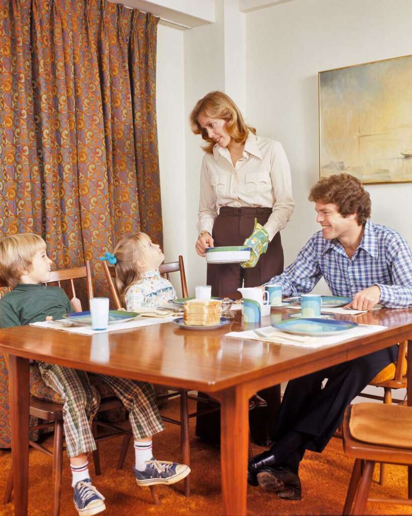 A family of four sits around a dining table. A woman is pouring juice. Two children sit on one side, and a man in a blue checkered shirt is seated on the other. The room has patterned curtains and a painting on the wall.