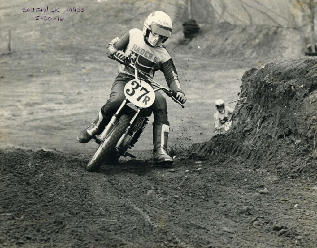 A motocross rider wearing protective gear and a helmet races a dirt bike on a rugged track. The bike has the number 37R on the front. Another rider is visible in the background. The setting is an outdoor, dirt-filled course.