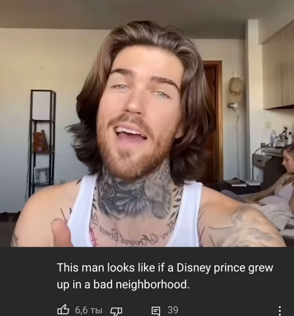 Man with long hair and tattoos wearing a white tank top. He is sitting indoors with a woman in the background. A comment below reads, "This man looks like if a Disney prince grew up in a bad neighborhood.