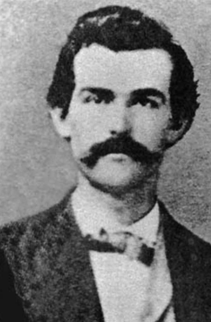 A black-and-white vintage portrait of a man with a prominent mustache wearing a suit and bow tie. The image appears old and has a grainy texture, typical of 19th-century photography.