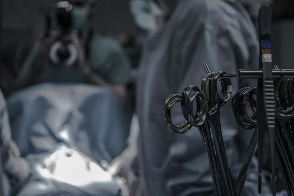 A dimly lit surgical room with instruments, such as scissors and clamps, hanging on a rack in the foreground. In the background, two masked figures in surgical attire stand near a patient on the operating table. One person is holding a camera.