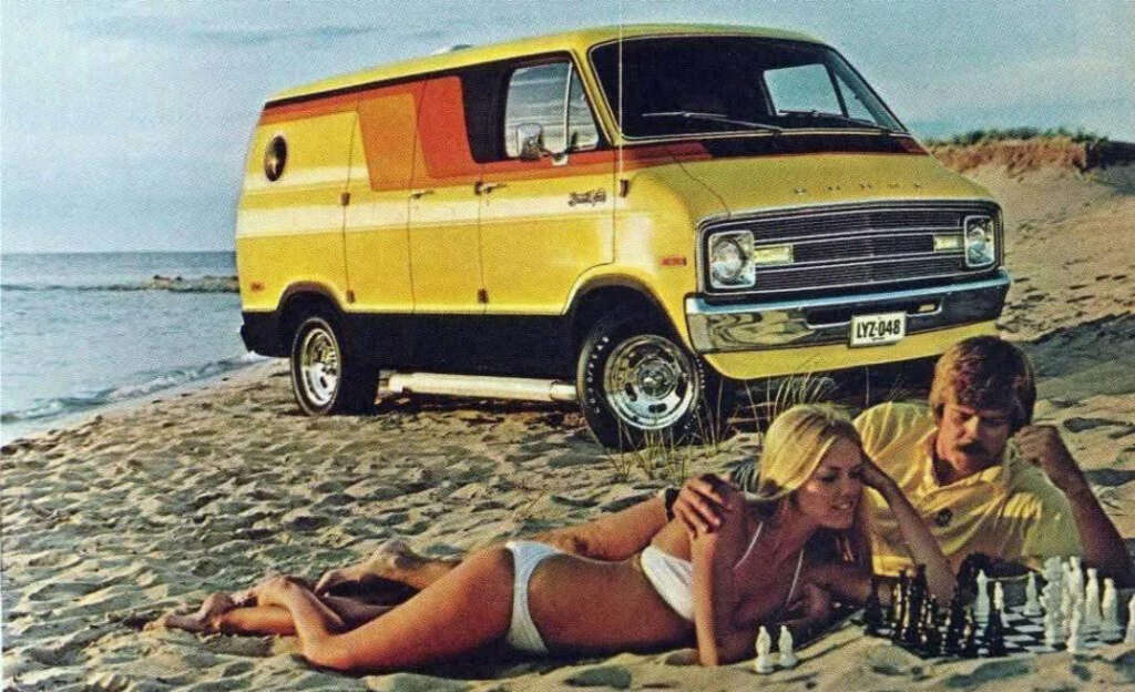 A yellow and orange van is parked on a beach. A man and woman are lying on the sand playing chess. The ocean is visible in the background under a clear sky.