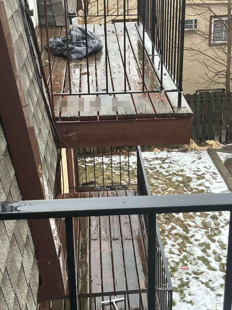 View of building balconies with wet wooden floors and metal railings. A garbage bag is visible on the top balcony. The ground below is partially covered in snow, and a wooden fence separates the property from neighboring buildings.