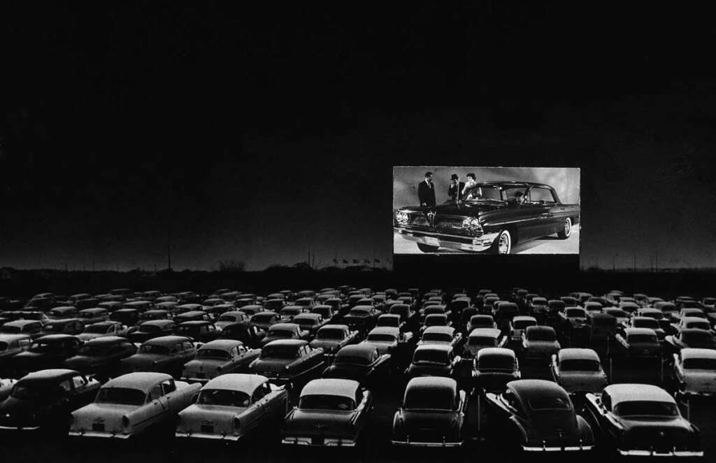 A black-and-white photograph of a crowded drive-in theater at night. Cars are parked and facing a large screen showing an image of a classic car with three people. The dark sky contrasts with the illuminated screen.