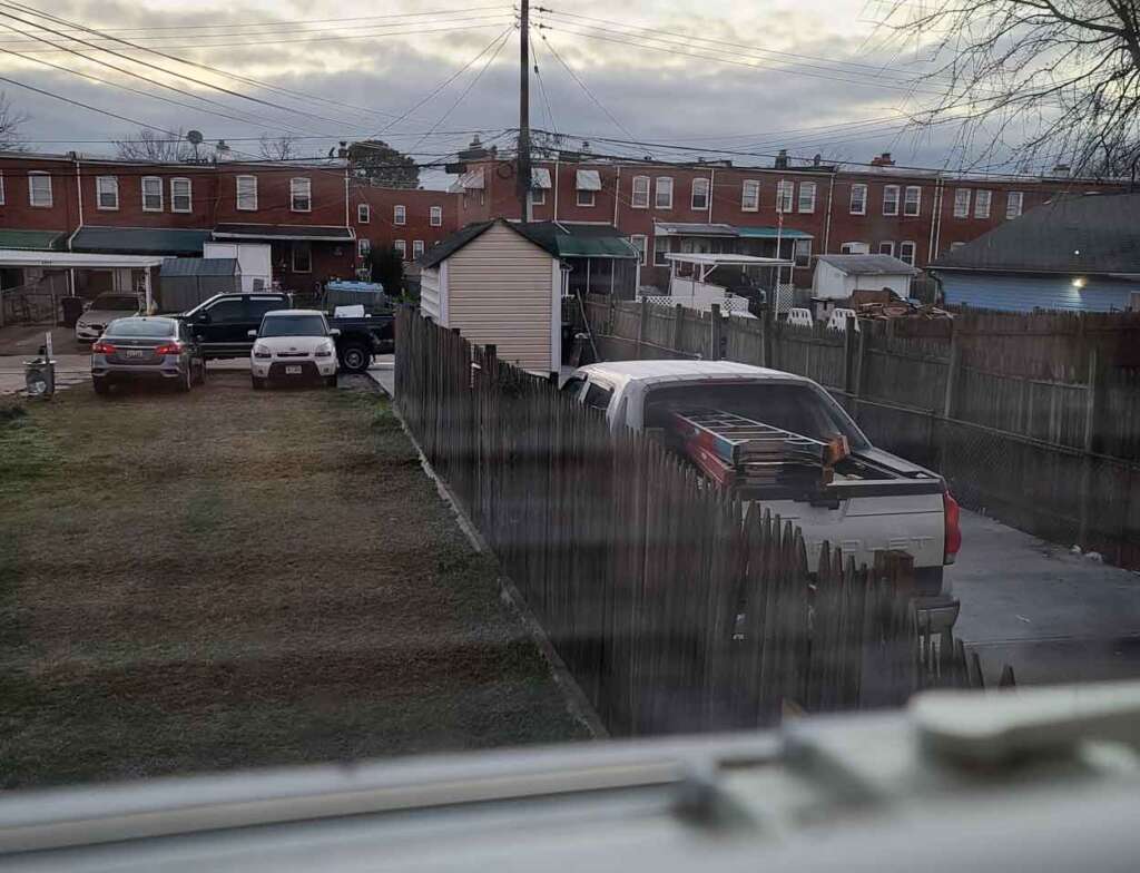 View from a window overlooking a backyard with parked cars, a pickup truck with items in the bed, and a row of brick houses in the background under a cloudy sky. A wooden fence separates the yards.