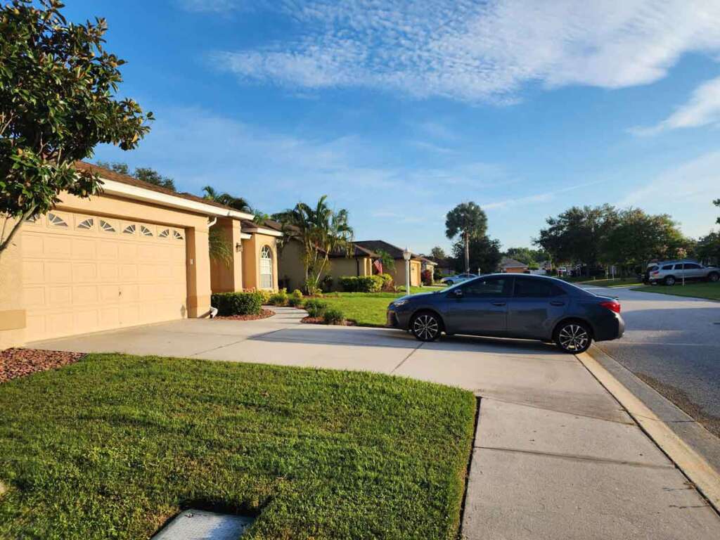 A suburban street with a one-story tan house and a well-kept lawn. A gray car is parked in the driveway. The sky is clear with a few clouds, and there are trees and shrubs lining the street.