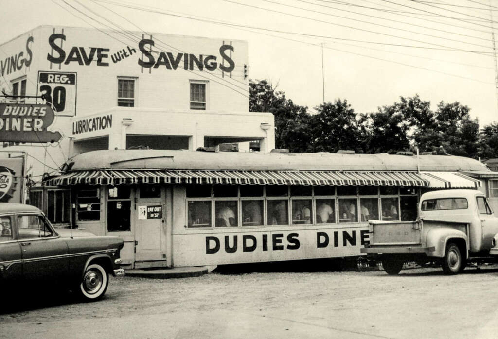 Black and white photo of Dudie's Diner, an old-fashioned diner converted from a railroad car, with a striped awning. Vintage cars and a truck are parked nearby. A building in the background has a "Save with Savings" sign.