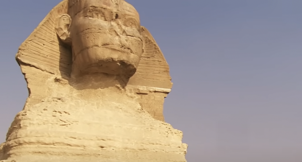 A close-up view of the Great Sphinx of Giza, showing its weathered limestone face and headdress against a clear blue sky. The Sphinx's body is partially visible, highlighting its ancient, carved texture.