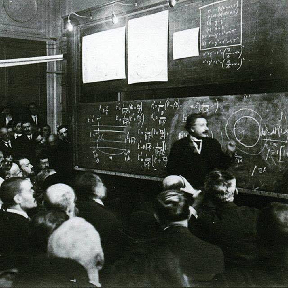 A man in a suit stands by a blackboard covered with complex equations and diagrams, addressing a seated audience in a lecture hall. The room is filled with attentive listeners, and additional charts are pinned above the board.