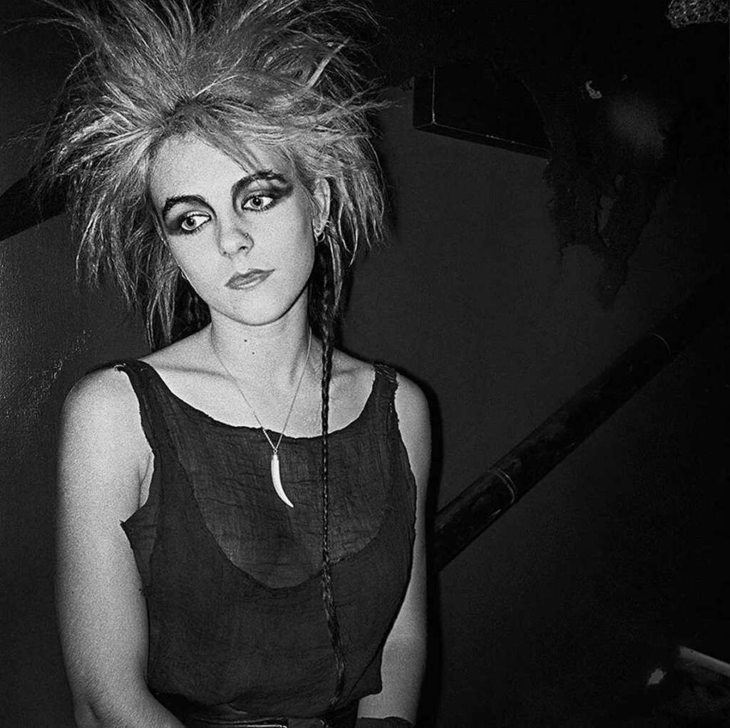 Black and white image of a person with spiky hair and dark eye makeup, wearing a sleeveless top and a necklace with a pendant. They are sitting against a dark background, looking to the side.