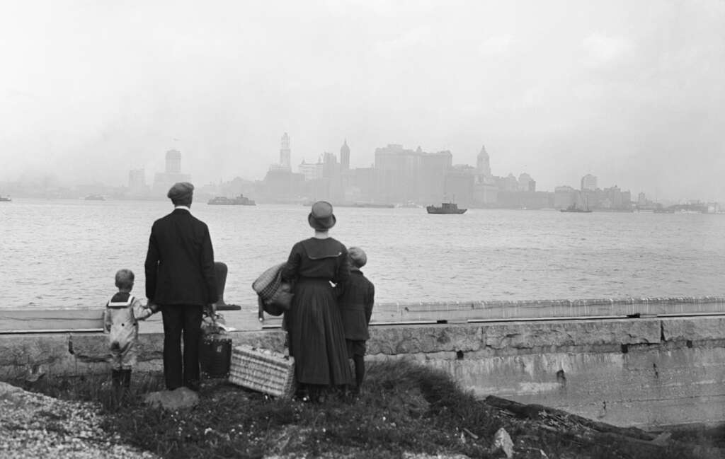 A black and white photo of a family standing by a waterfront with luggage, gazing at the distant skyline of New York City shrouded in fog. The family includes a man, a woman, and three children.