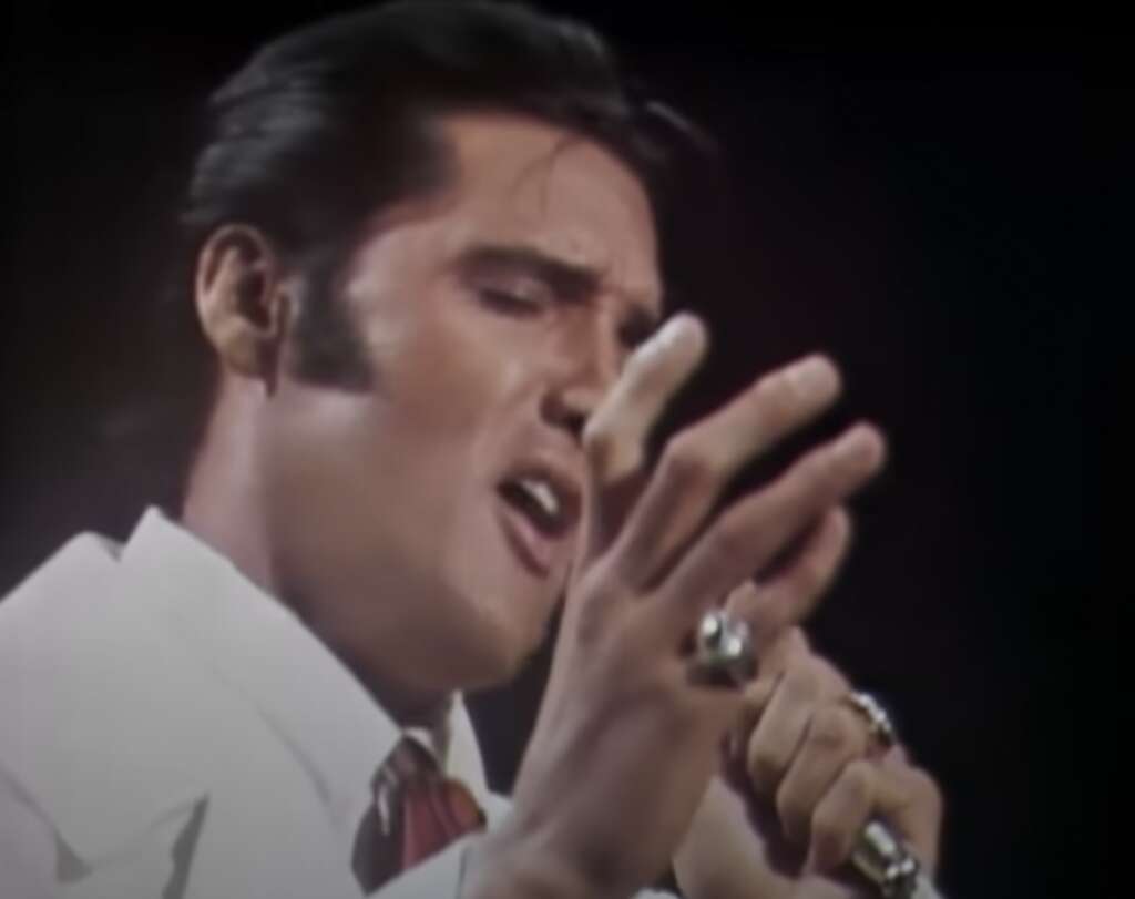 A person with dark hair sings passionately into a microphone, wearing a white outfit and rings, against a dark background.