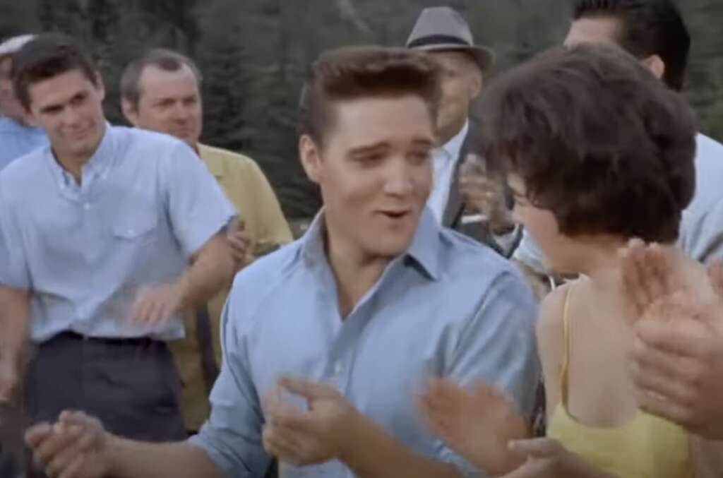 A group of people outdoors, smiling and clapping. A man in a blue shirt is gesturing and looking at a woman in a yellow dress. Trees are visible in the background.