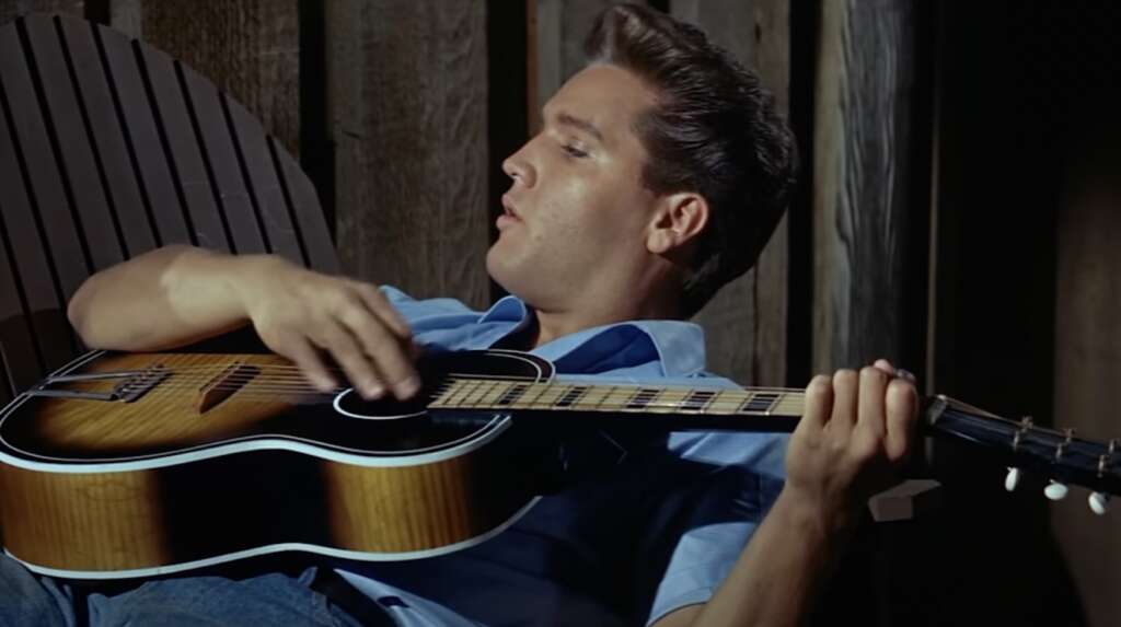 A person reclining in a wooden chair strums an acoustic guitar. They are wearing a light blue shirt and have styled, dark hair. The background consists of wooden planks.