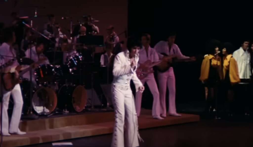 A singer in a white jumpsuit performs on stage. A band with a drummer and guitarists in white suits and backup singers in black outfits with yellow capes accompany. The background is dimly lit, focusing on the lively performance.