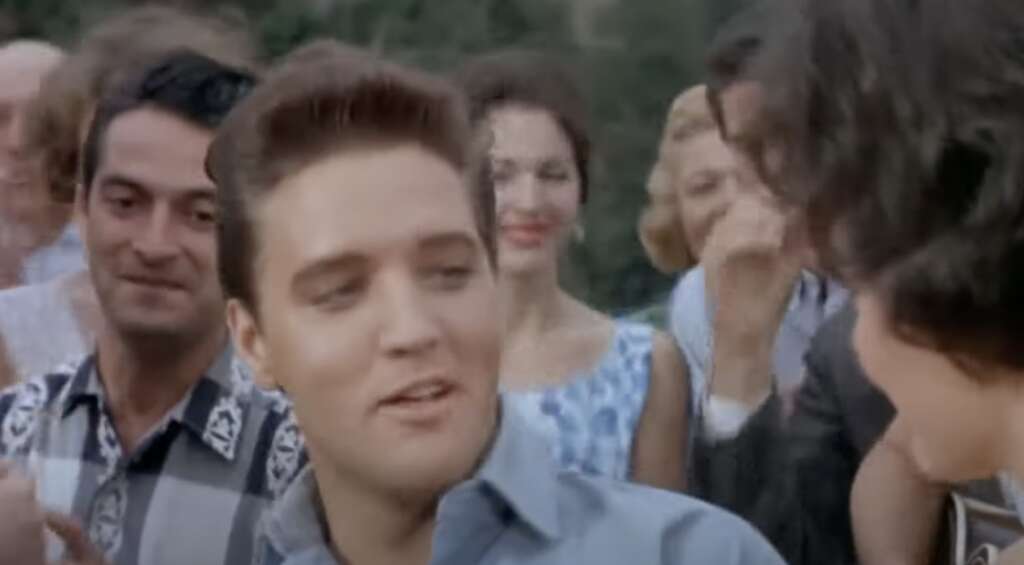 A group of people are gathered outdoors. The focus is on a young man with styled dark hair in a light shirt, smiling slightly. Other people in the background are smiling and engaged in conversation.