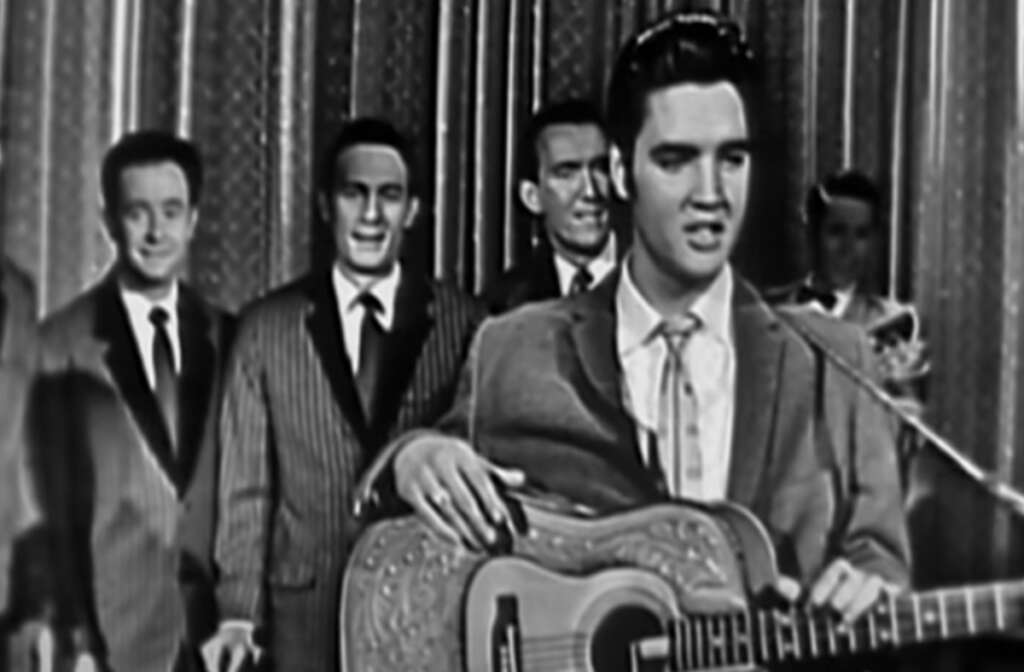 A black-and-white photo of a man with dark hair standing in front, holding a guitar. Four other men are in the background, smiling and dressed in suits, standing in front of a curtain.