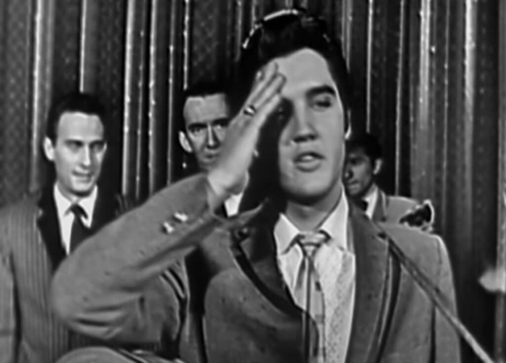 A black-and-white photo of a singer on stage, wearing a suit and tie, saluting. He is in front of a microphone, with backup band members visible in the background. The setting has curtains and an old-fashioned microphone stand.