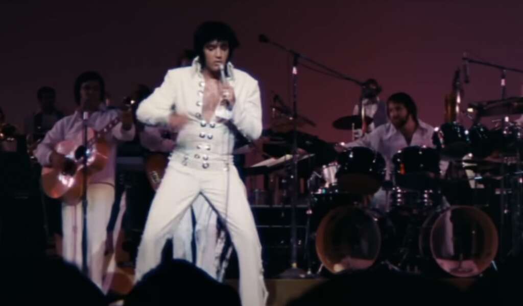 A performer in a white, embellished jumpsuit sings and gestures energetically on stage. Behind him, a band is playing various instruments. The background is lit with warm colors.