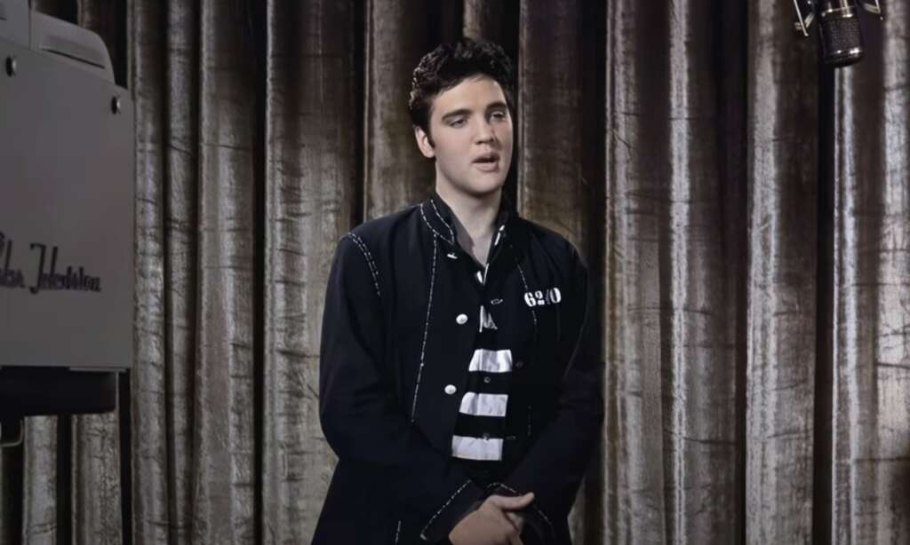 A man with dark hair and an embroidered jacket is standing in front of a curtain, holding his hands together. A vintage camera is partially visible to the left. The setting has a retro style, suggesting a performance or recording session.