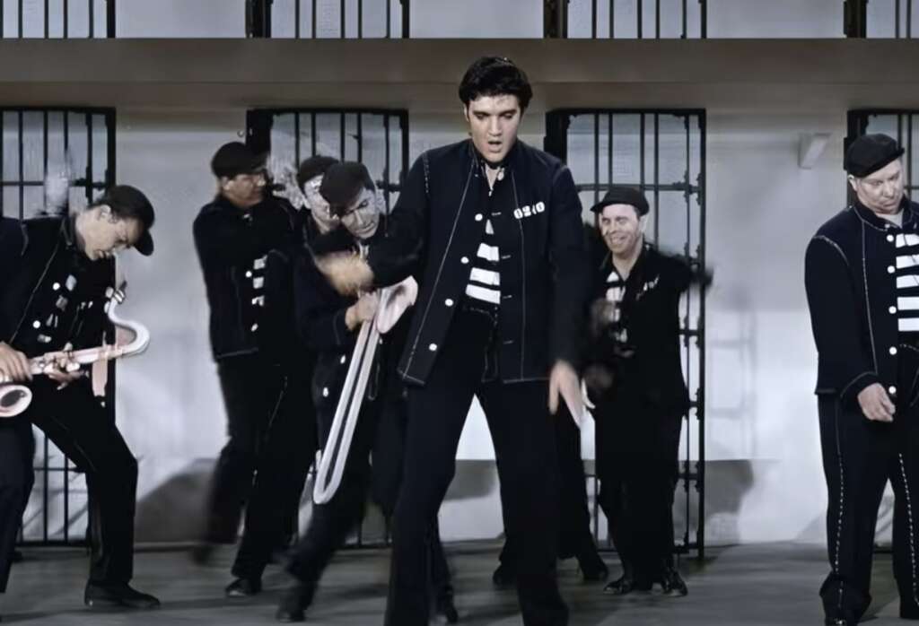 A group of men in black prison uniforms perform a dance routine in front of barred cell doors. The central figure energetically swings a rope while others pose or dance around him. They wear matching black caps and outfits with white stripes.