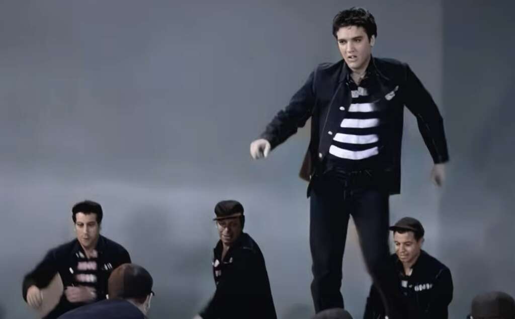 A man wearing a striped shirt and dark jacket dances energetically on a raised platform. Three other men in similar outfits are nearby, also engaged in dancing. The background is plain and gray, emphasizing their movements.