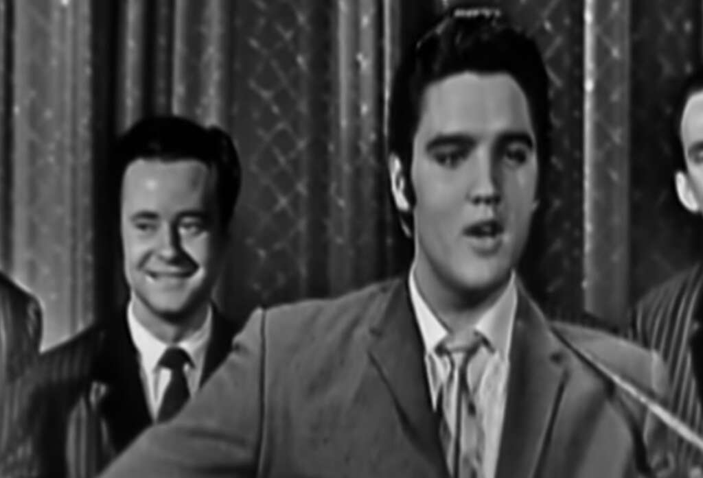 A black and white image of a male musician playing a guitar and singing into a microphone. He is dressed in a suit and tie. A smiling man in a suit stands behind him, with patterned curtains in the background.