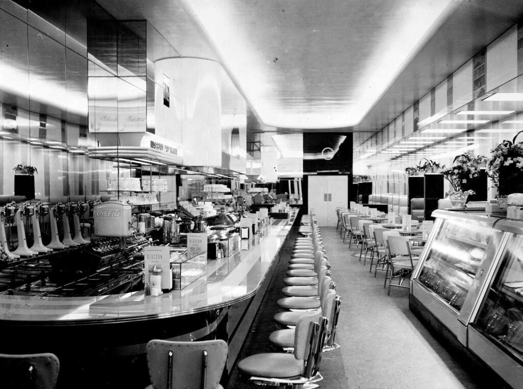 Black and white photo of a vintage diner interior with a long counter and row of stools on the left. Opposite, tables with chairs line the wall. Retro decor includes a soda fountain and display case. Overhead lighting brightens the scene.