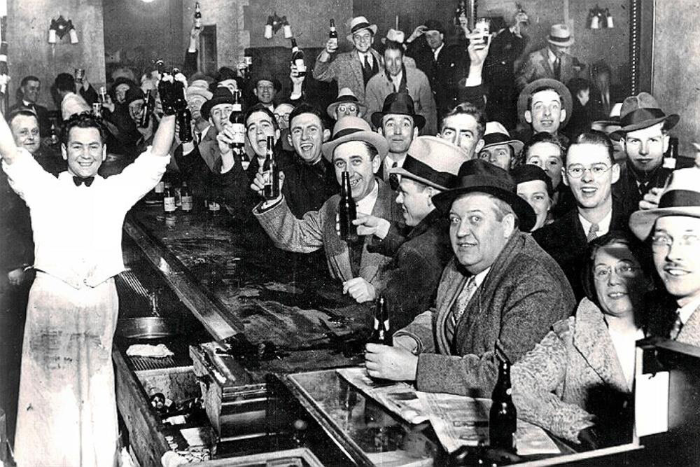 A crowded bar scene from the Prohibition era, with men and women in 1920s attire, holding up drinks and smiling. A bartender stands at the bar, arms raised in celebration. The setting is lively and jubilant.