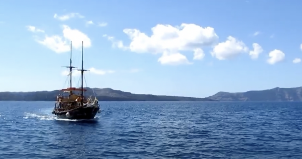 A small sailboat with two masts sails on a calm, blue sea under a clear sky with scattered clouds. In the background, there are distant hills or mountains along the horizon.