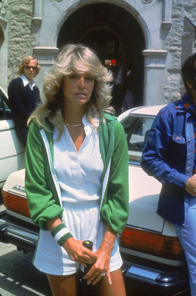 Person with long hair, wearing a white outfit and green jacket, stands in front of a car. They are holding a tennis racket. Another person in a blue jacket and a person with sunglasses are in the background near a stone building.