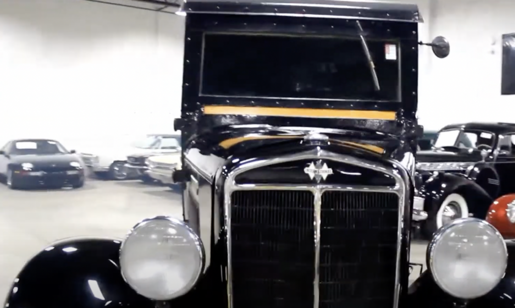 A vintage black car with a wide front grille and large round headlights is parked in an indoor showroom. Behind it, several other classic cars are visible. The lighting highlights the shiny surface of the car.