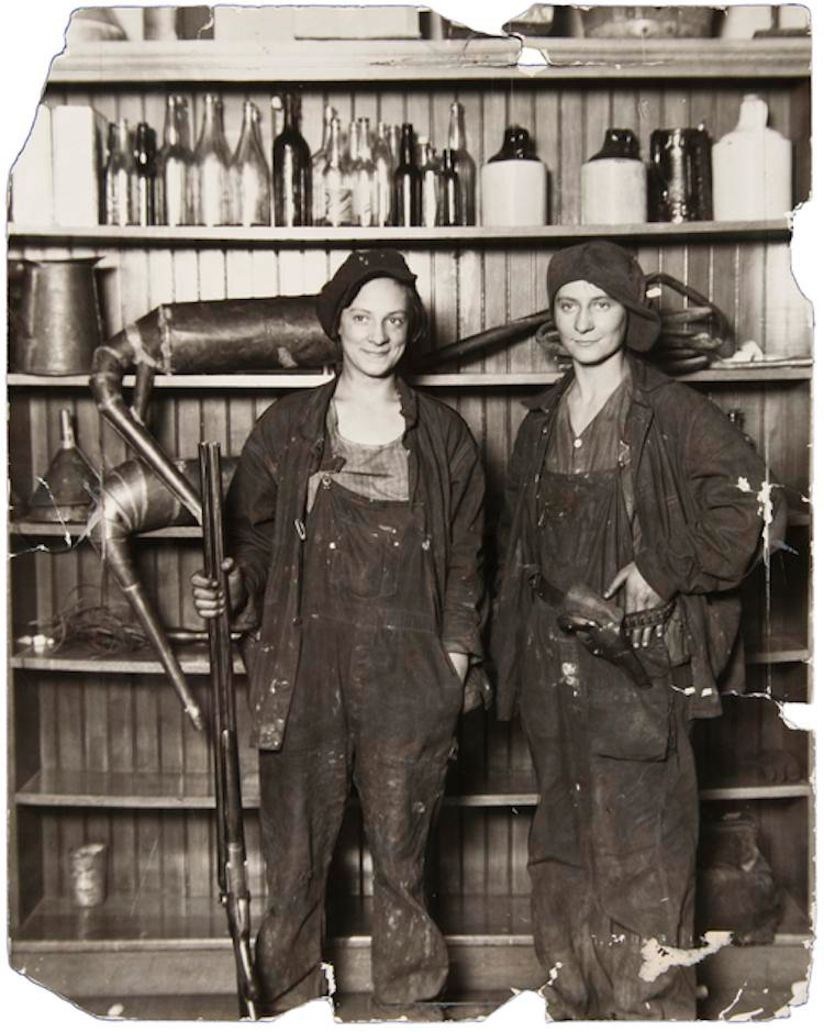 Two people in work attire stand in front of shelves filled with bottles and jars. They are holding tools and wearing caps and overalls, suggesting a workshop or industrial setting. The image has a vintage, worn appearance.