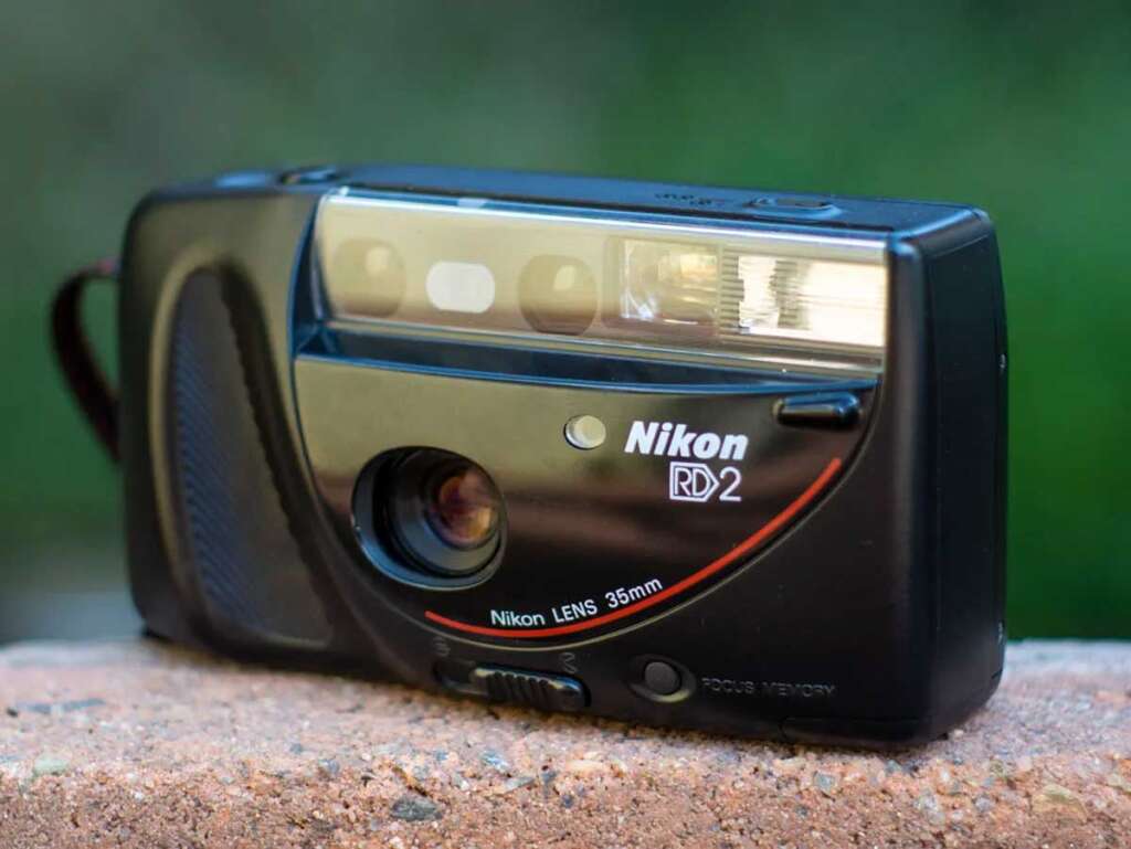 A black Nikon RD2 35mm film camera sits on a textured surface. The camera has a visible lens and flash on the front, with brand markings in white and red. The background is a blurred green.