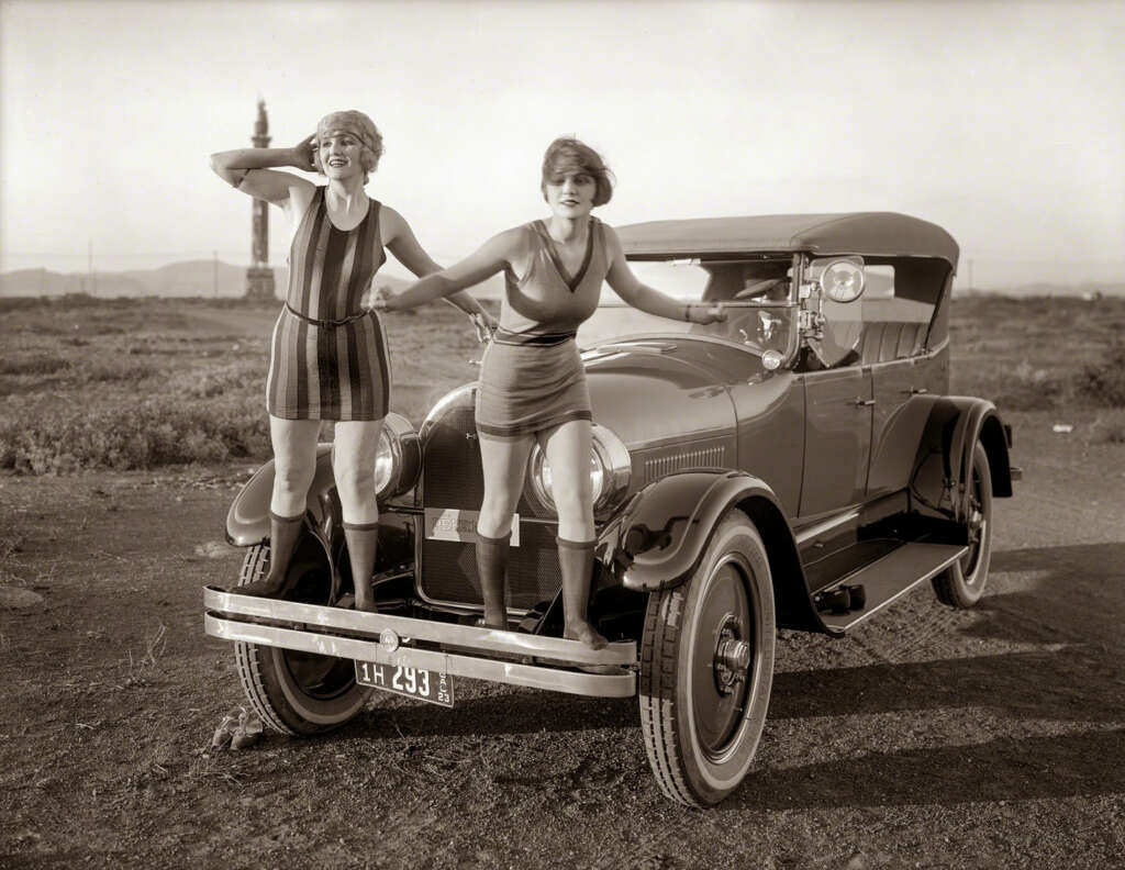 Two women in 1920s swimsuits stand on the bumper of a vintage convertible car parked on a dirt road. One stands with hands on her head, and the other holds a small towel, with a distant structure visible in the background.