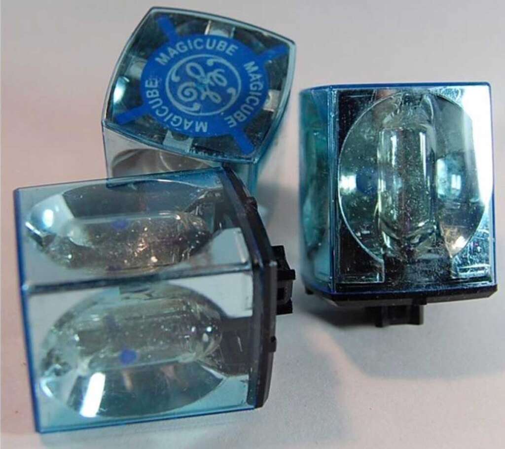 Three vintage GE Magicube flashbulbs, one upright, two tilted, with visible reflective surfaces and a blue and white logo on top. They have rectangular shapes with blue-tinted casings.