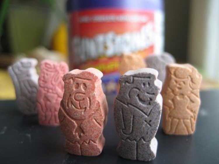 Close-up of Flintstones themed chewable vitamins shaped like cartoon characters, with a container of vitamins blurred in the background. The vitamins are in various colors, including pink, gray, and brown.